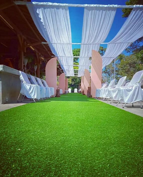 Area terbuka UTC beratap kain putih dengan karpet rumput sintetis dan deretan kursi bersarung putih, ditata untuk sebuah acara.