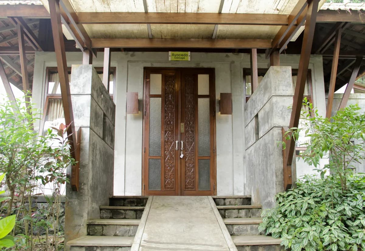 Pintu masuk Avocado Cottage dengan daun pintu kayu berukir, tangga batu, dan tanaman hijau di kedua sisinya.