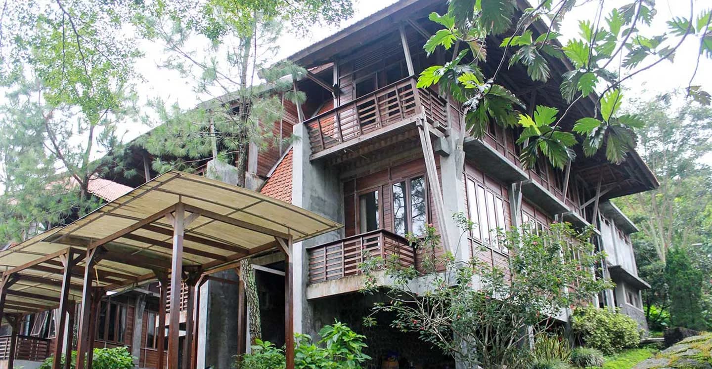 Bangunan dua lantai Cassava & Durian Cottage berdinding kayu dengan balkon, dikelilingi pohon pinus.