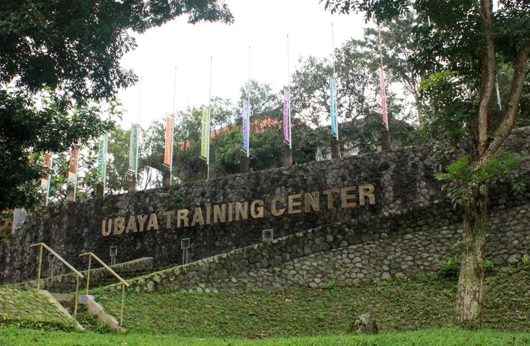 Dinding batu bertuliskan Ubaya Training Center di tepi jalan kawasan, dengan deretan umbul-umbul dan pepohonan di atasnya.