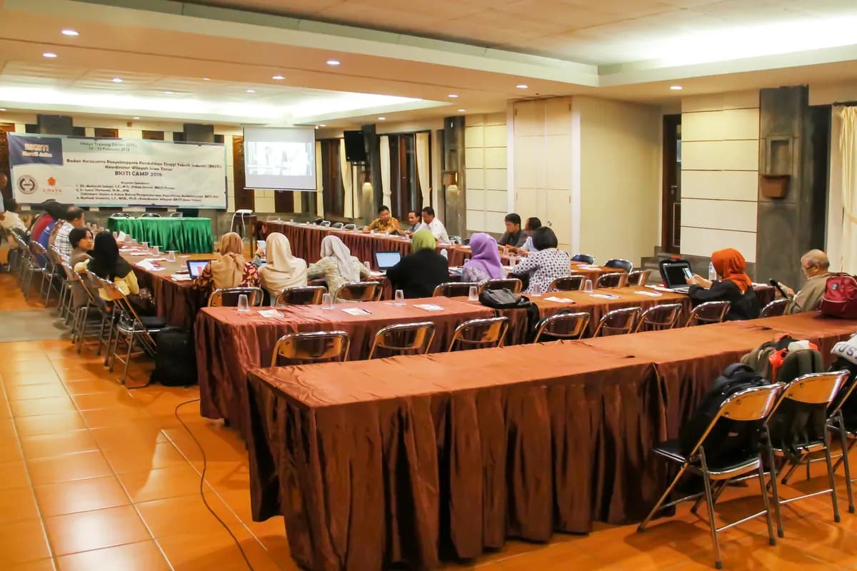 Hall UTC ditata gaya konferensi dengan meja panjang berskirting dan layar proyektor, peserta sedang mengikuti sesi.