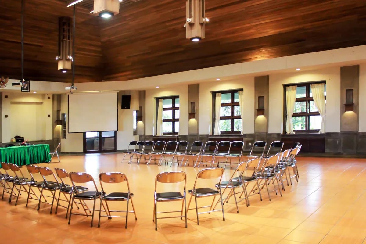 Interior Multifunction Hall UTC dengan langit-langit kayu, deretan kursi lipat, dan layar proyektor di depan ruangan.