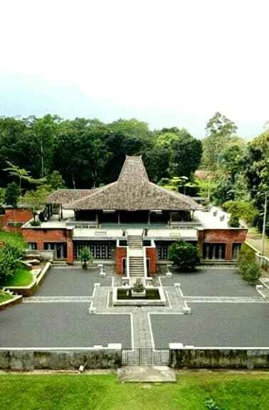 Pendopo Mojopahit beratap joglo dilihat dari kejauhan, dengan halaman berpaving, kolam kecil, dan tangga di depannya.