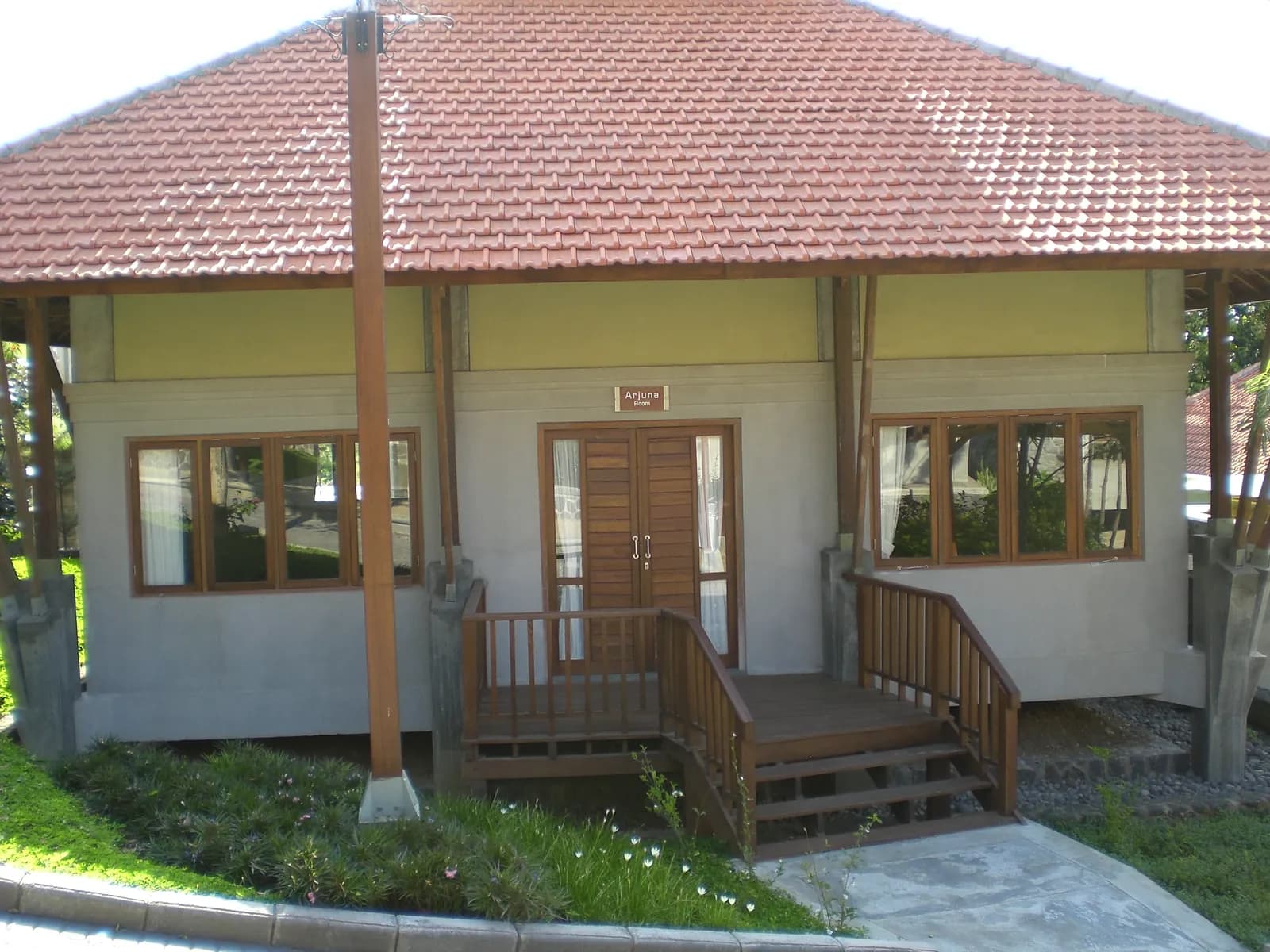 Bangunan Ruang Arjuna dari luar dengan papan nama Arjuna Room, atap genteng, teras kayu berpagar, dan taman di depannya.